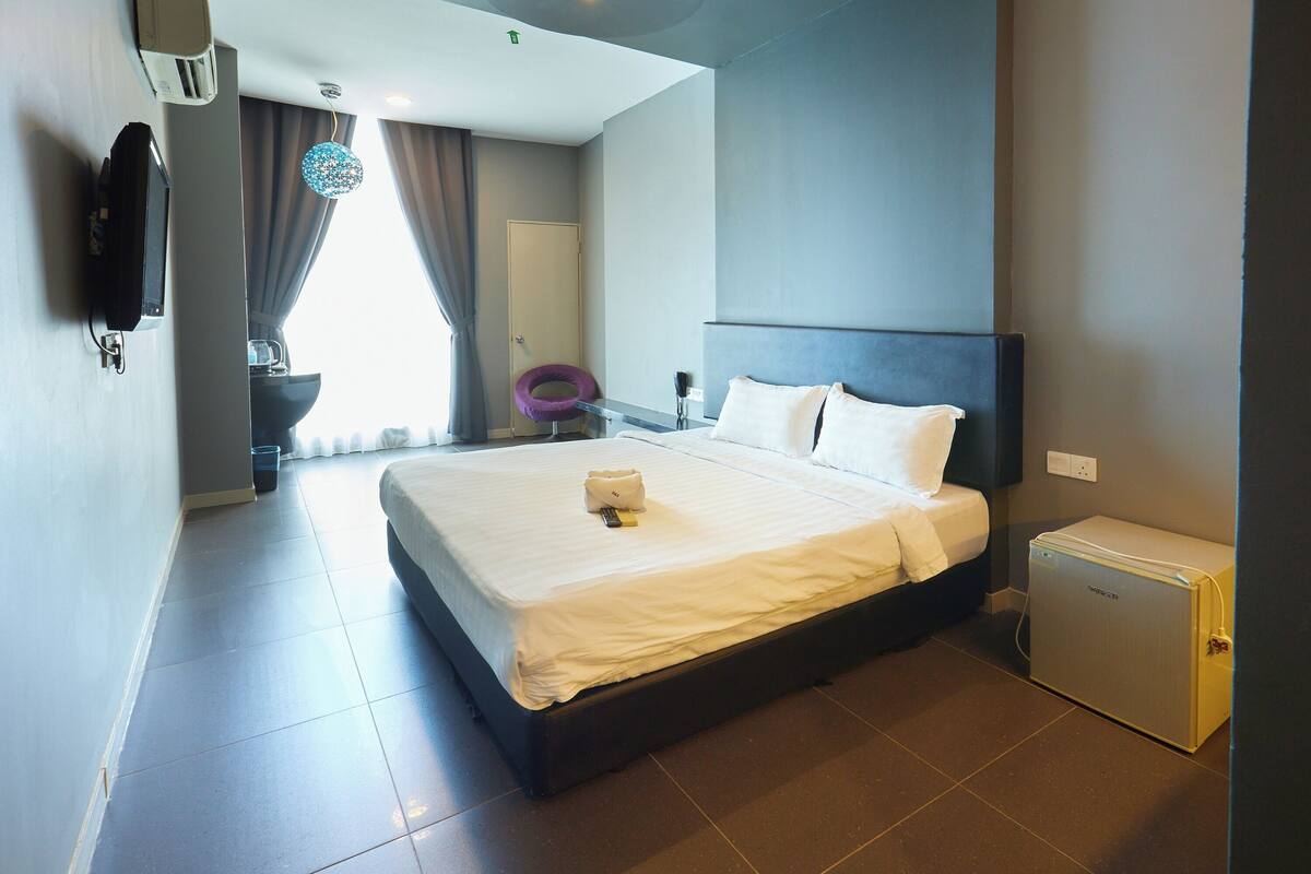 Une chambre d’hôtel moderne dotée d’un grand lit, d’un petit réfrigérateur, d’un bureau avec une chaise, d’une télévision et d’une fenêtre avec des rideaux.