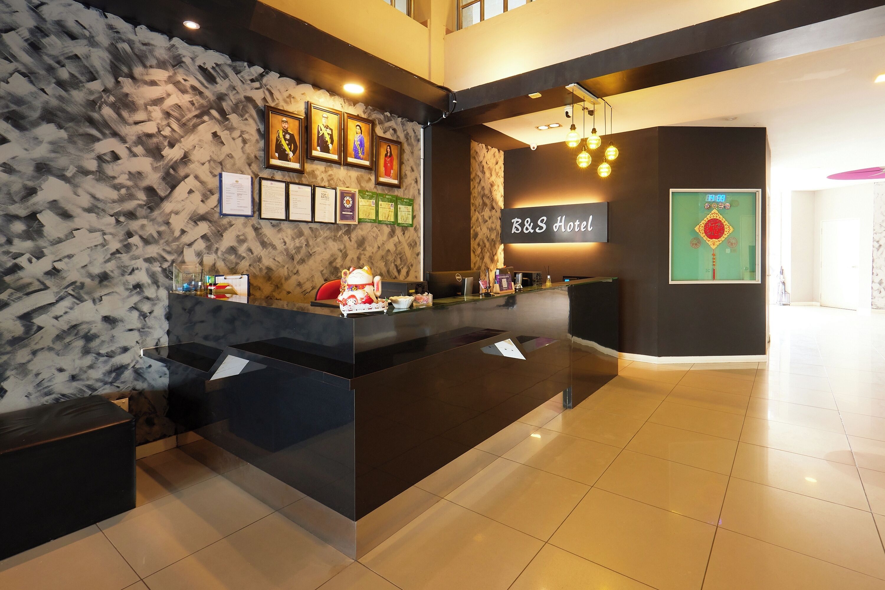 Zona d'entrada d'un hotel amb un disseny modern, que inclou una recepció, diplomes emmarcades i un rellotge digital.