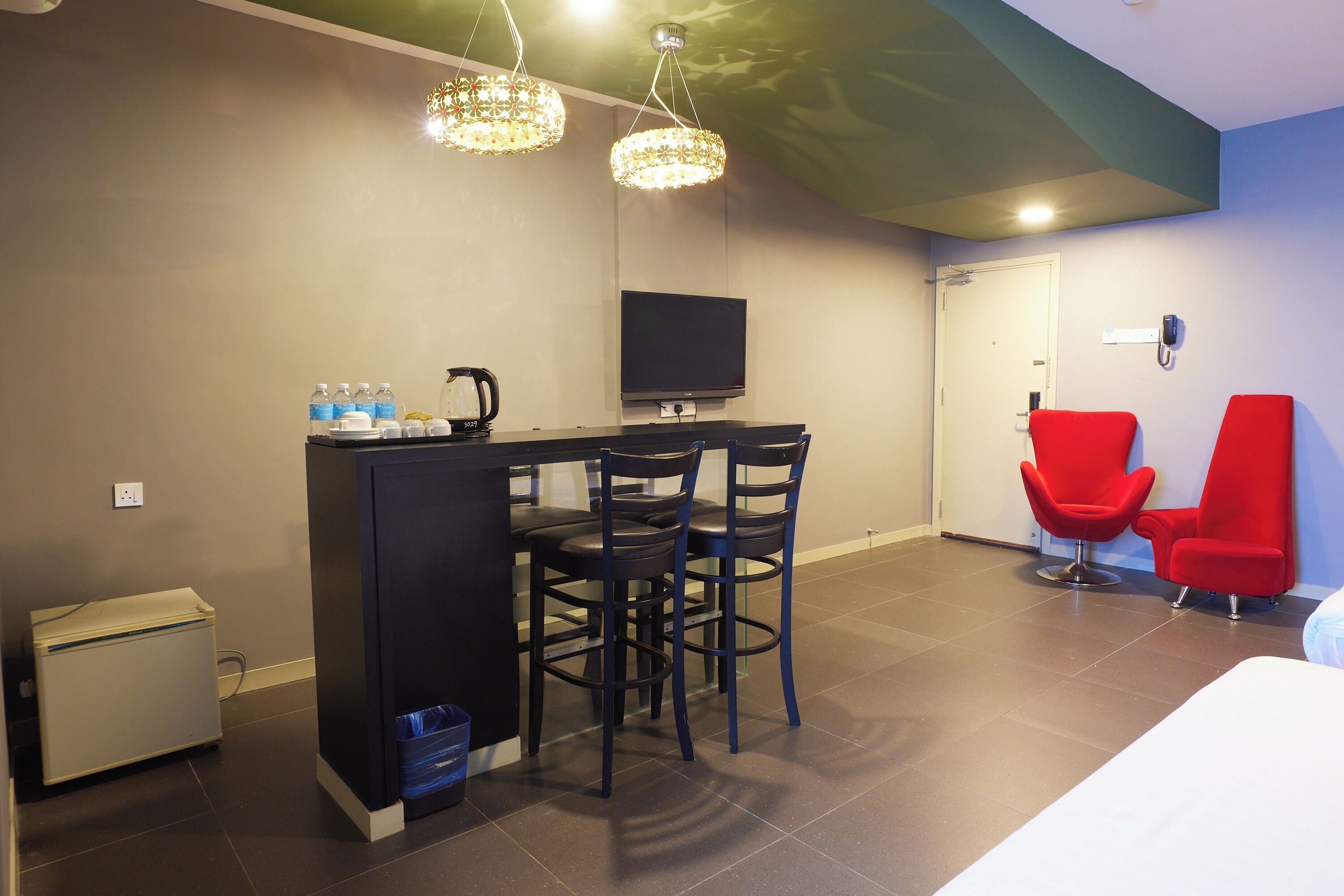 A modern hotel room with a kitchenette, a flat-screen TV, and two red chairs.