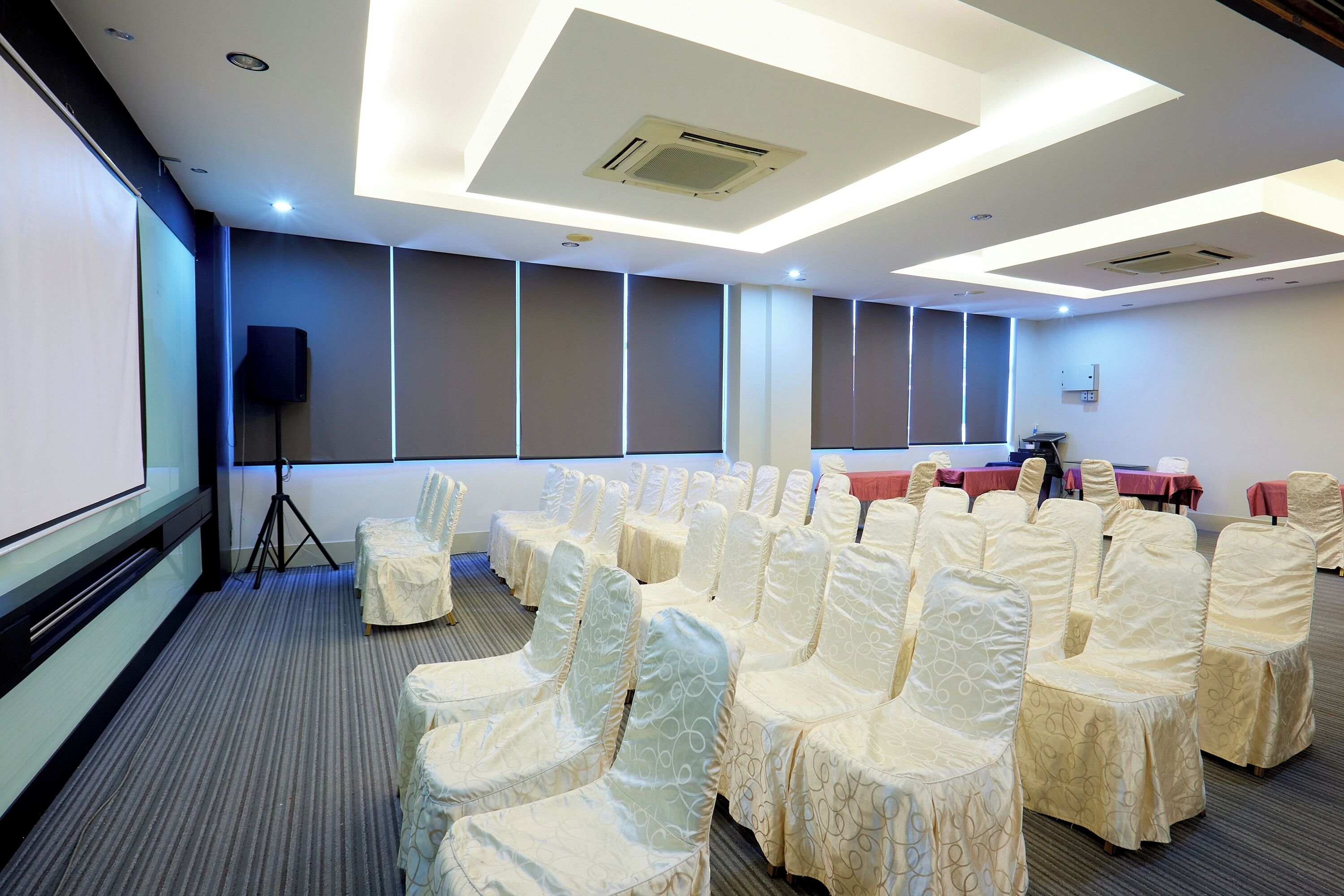 Une salle de conférence avec des rangées de chaises recouvertes d’un tissu blanc, un écran de projection et des tables recouvertes de nappes rouges.