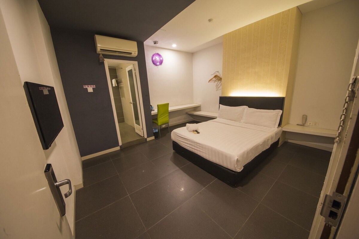 A hotel room with a bed, a desk, a chair, a wardrobe, and an air conditioning unit.