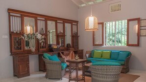A spacious living area with a large wooden cabinet, wicker furniture, and a view of greenery through the window. - Bungalow Sud (Koggala)