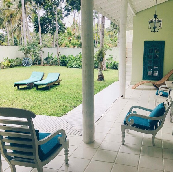 A covered patio area with two lounge chairs and a table, surrounded by a lush garden with palm trees and a white fence. - Bungalow Sud (Koggala)