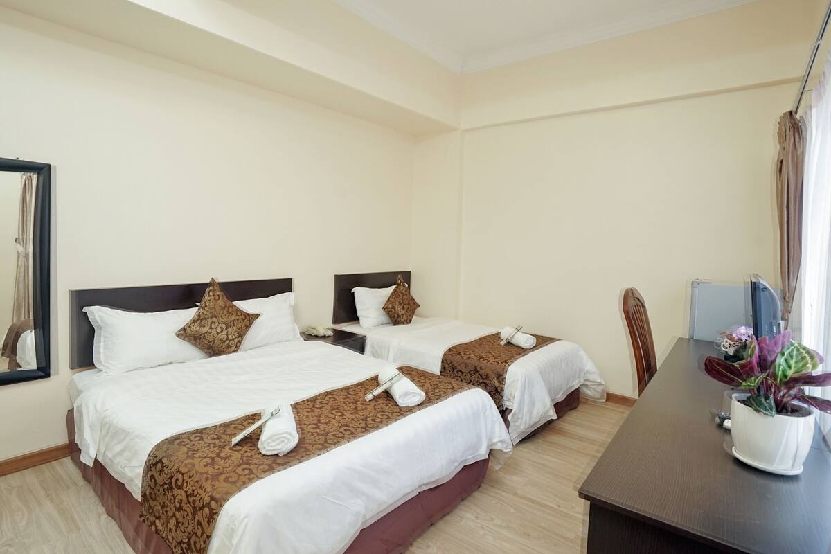 Quarto de hotel com duas camas, cabeceira de madeira, uma mesa de jantar e um vaso com planta.
