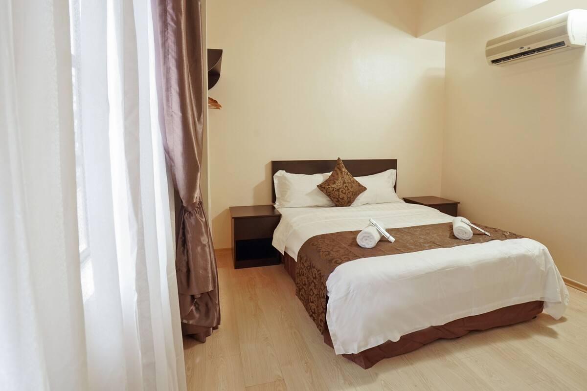 Cama bem arrumada com roupa de cama branca e marrom, um travesseiro e duas toalhas enroladas. Há uma mesa de cabeceira ao lado da cama e um aparelho de ar condicionado fixado na parede.