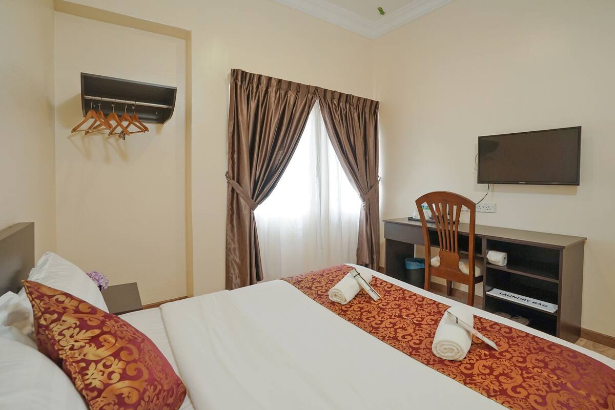 Quarto de hotel com cama, escrivaninha, cadeira, televisão e prateleira com toalhas.