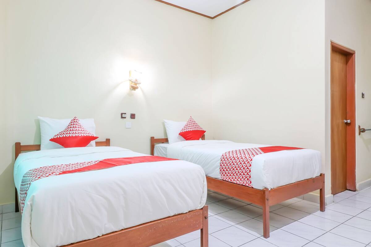A room with two wooden beds, white bedding, red pillows, and a wall-mounted light fixture.