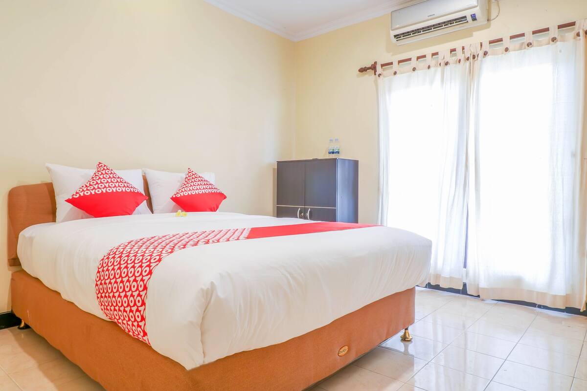A neatly made bed with a patterned bedspread and pillows, a wardrobe, and a large window with curtains.