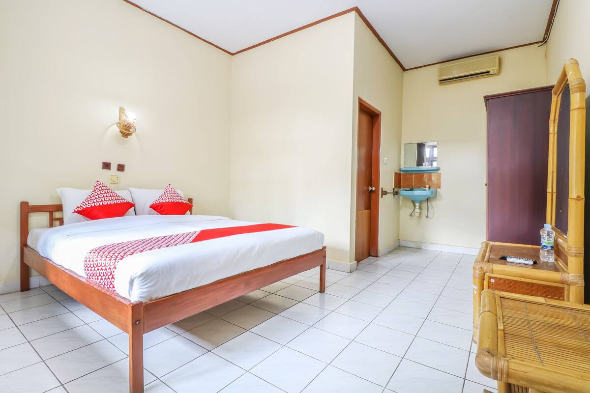 A bedroom with a wooden bed, red and white bedding, a bedside table with a remote control, a mirror, and a bathroom with a sink and mirror.