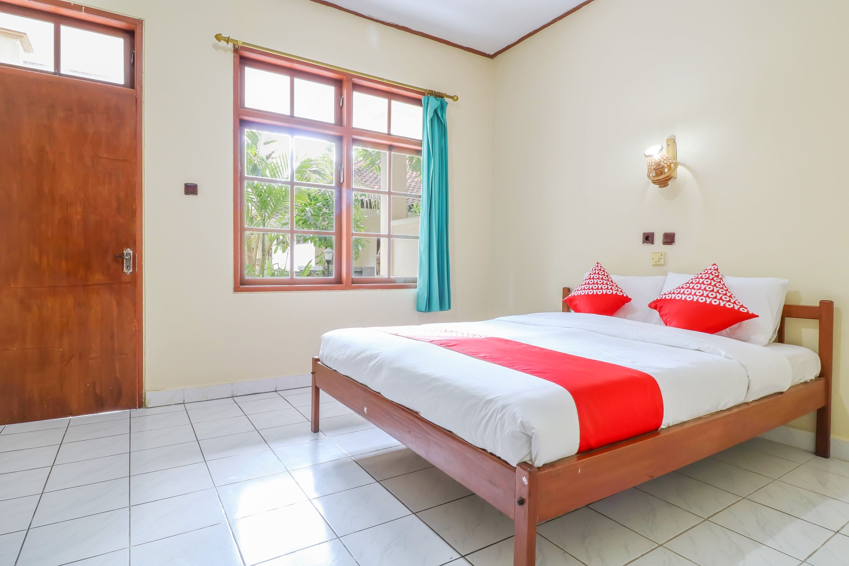 A bedroom with a wooden bed, white and red bedding, a window with a curtain, and a wall-mounted light fixture.
