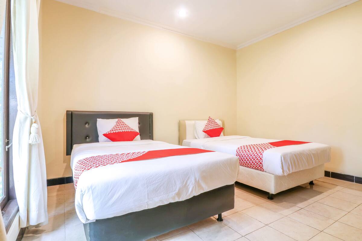A hotel room with two beds, each with a red and white patterned bedspread and matching pillows. The room has a neutral color scheme with cream walls and a tiled floor.