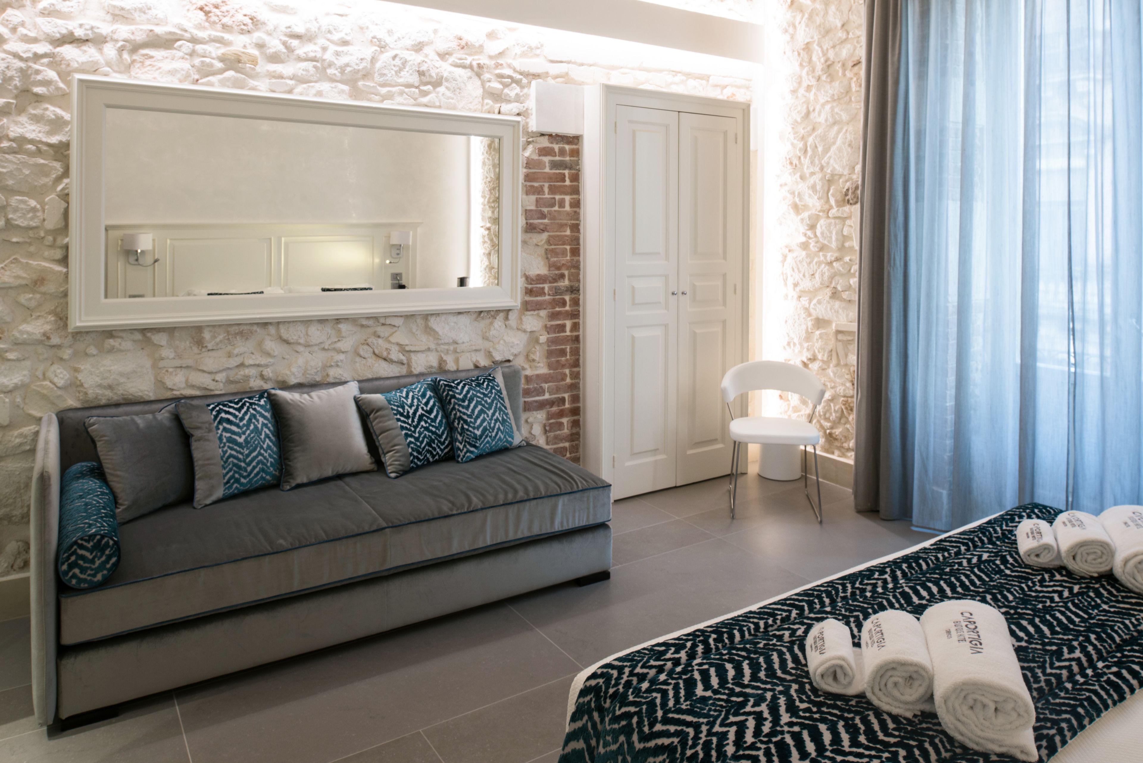 Une chambre moderne avec un mur en pierre, un canapé gris agrémenté de coussins à motifs, un grand lit recouvert d’une courtepointe à motifs, une chaise blanche et un grand miroir placé au-dessus du canapé.