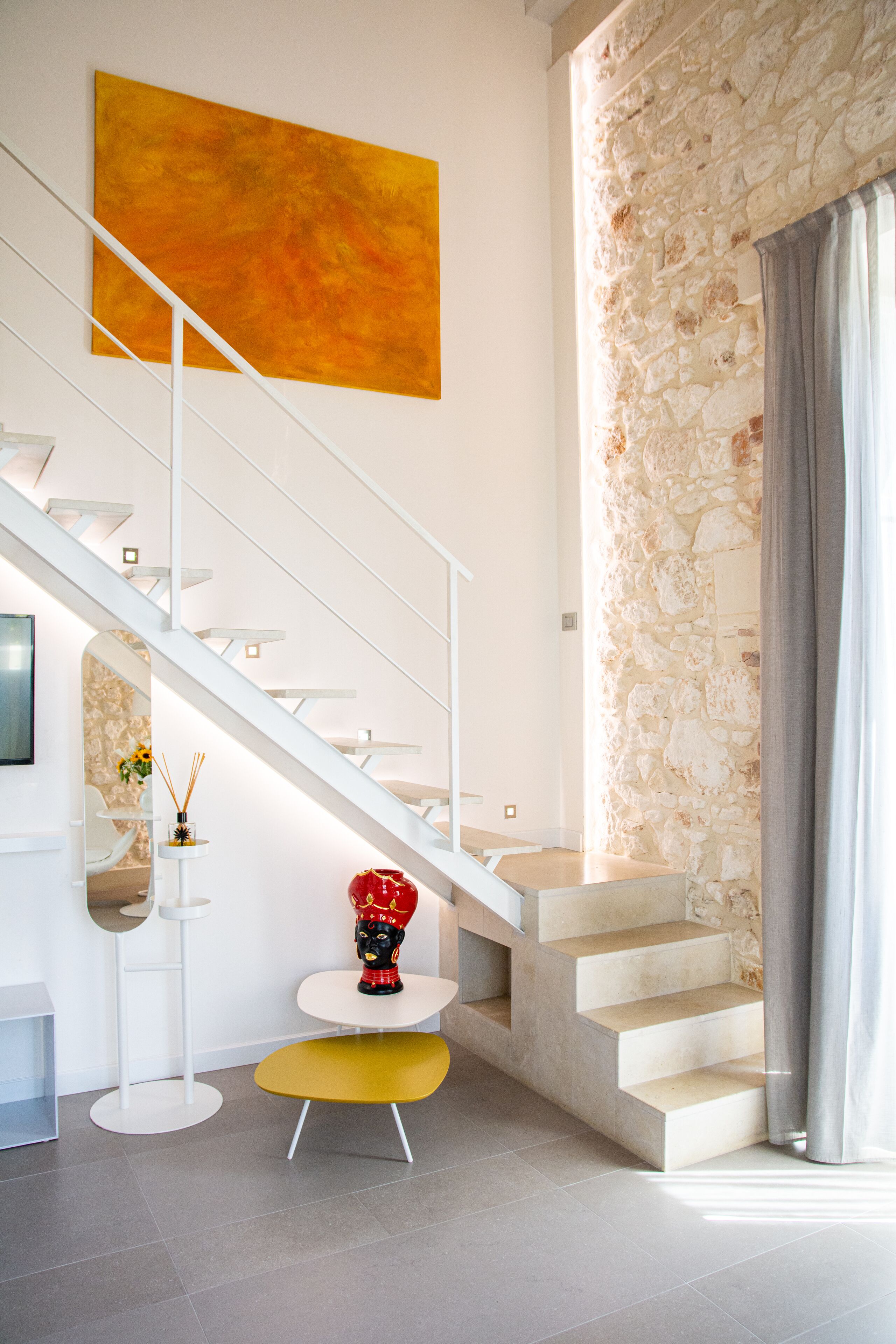 A modern interior with a staircase, a large abstract painting, a stone wall, and a small round table with a red vase.