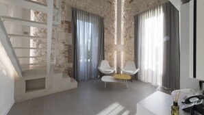 A modern hotel room with a stone wall, a staircase, a seating area with two chairs, a small table, and a desk with a phone and a lamp. - Caportigia Boutique Hotel (Syracuse)