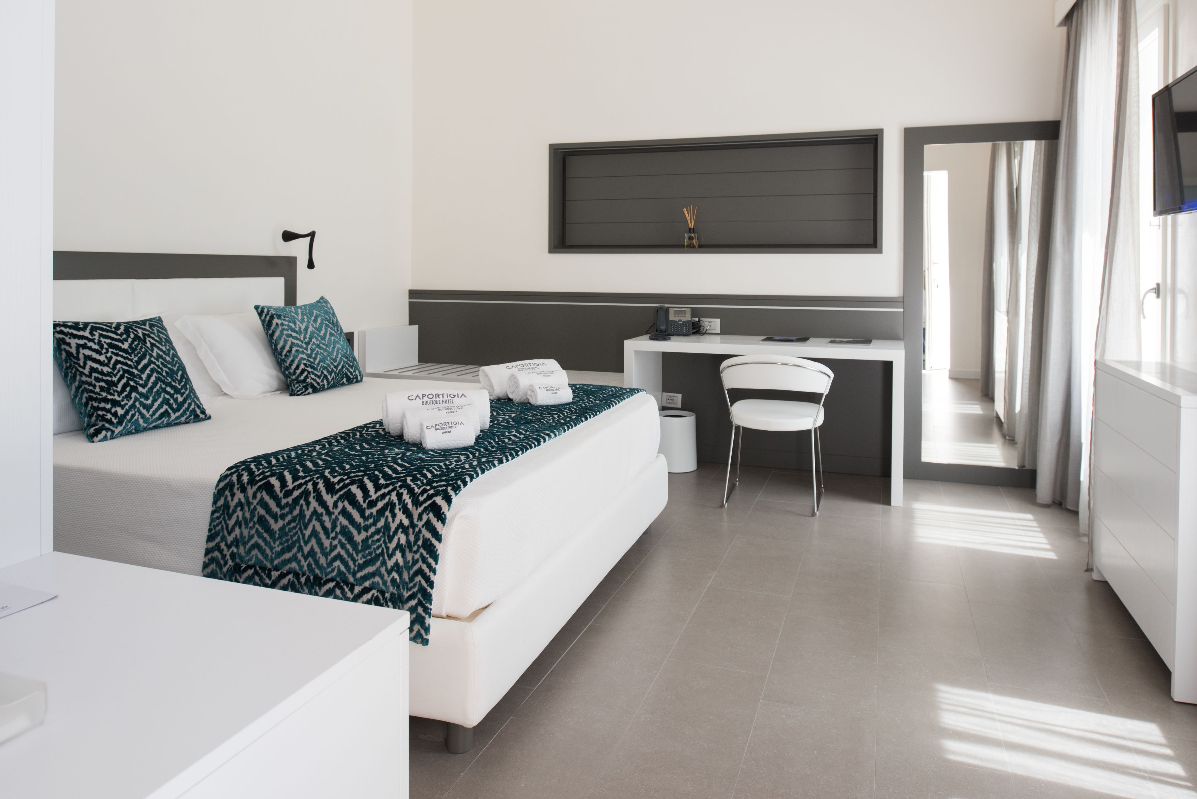 Une chambre d’hôtel moderne, équipée d’un lit, d’un bureau et d’une chaise. Un téléviseur à écran plat est fixé au mur, il y a une fenêtre avec des rideaux, et une serviette sur laquelle est inscrit le nom de l’hôtel.