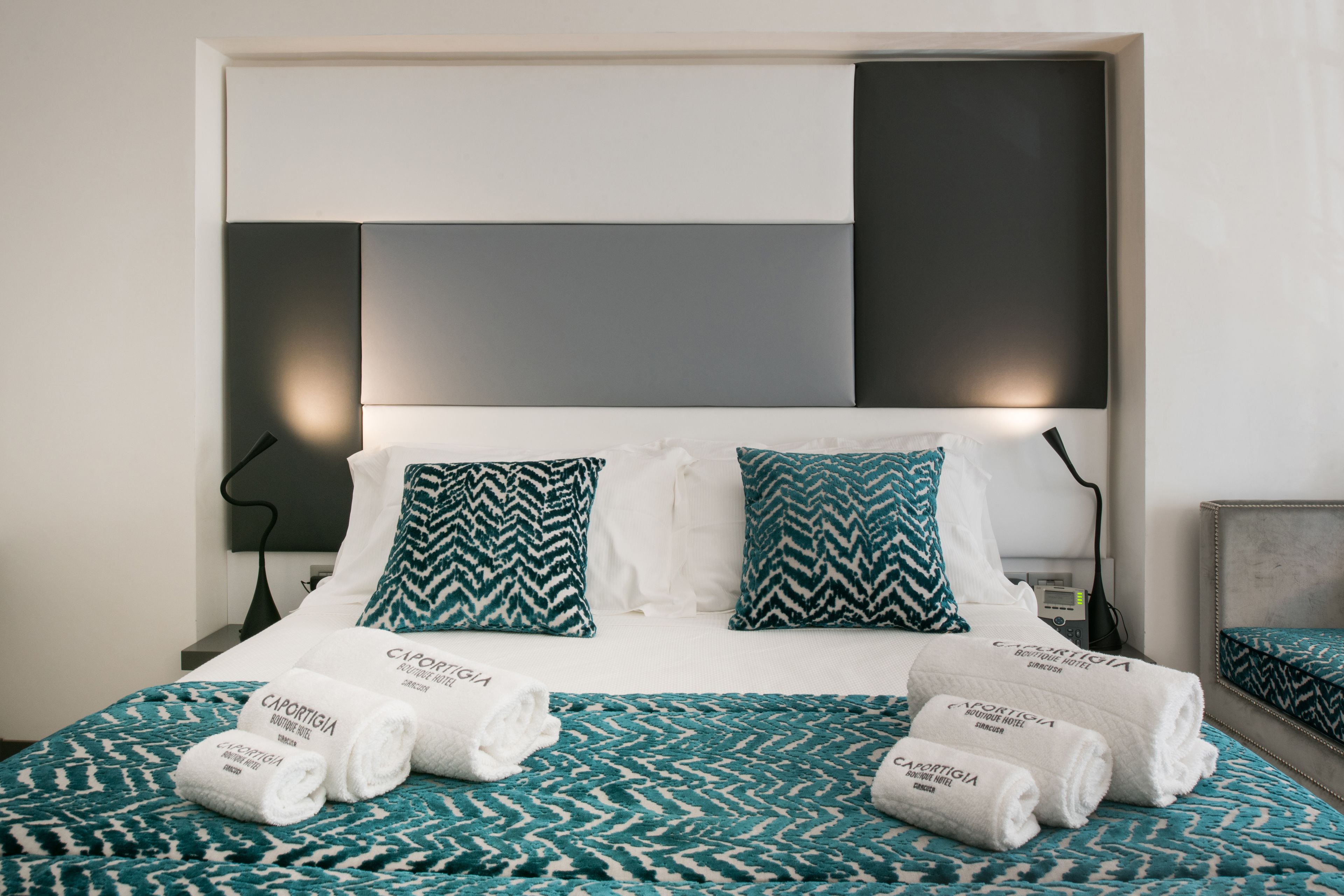 Een hotelkamer met een bed, beddengoed in wit en blauwgroen patroon, twee kussens en opgerolde handdoeken met tekst.