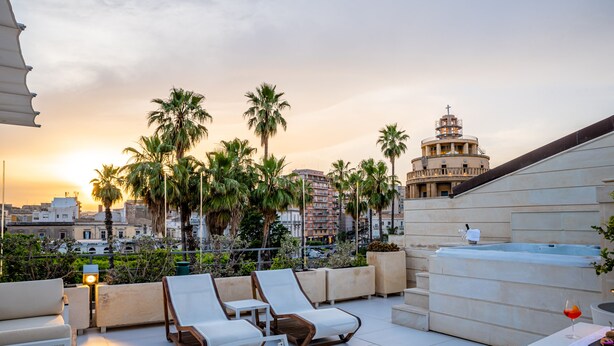 A rooftop terrace with lounge chairs, a hot tub, and a view of palm trees and buildings. - Caportigia Boutique Hotel (Syracuse)