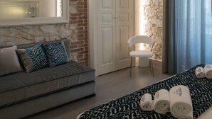 A modern hotel room with a stone accent wall, a grey sofa, a large mirror, a chandelier, and a neatly made bed with rolled towels. - Caportigia Boutique Hotel (Syracuse)