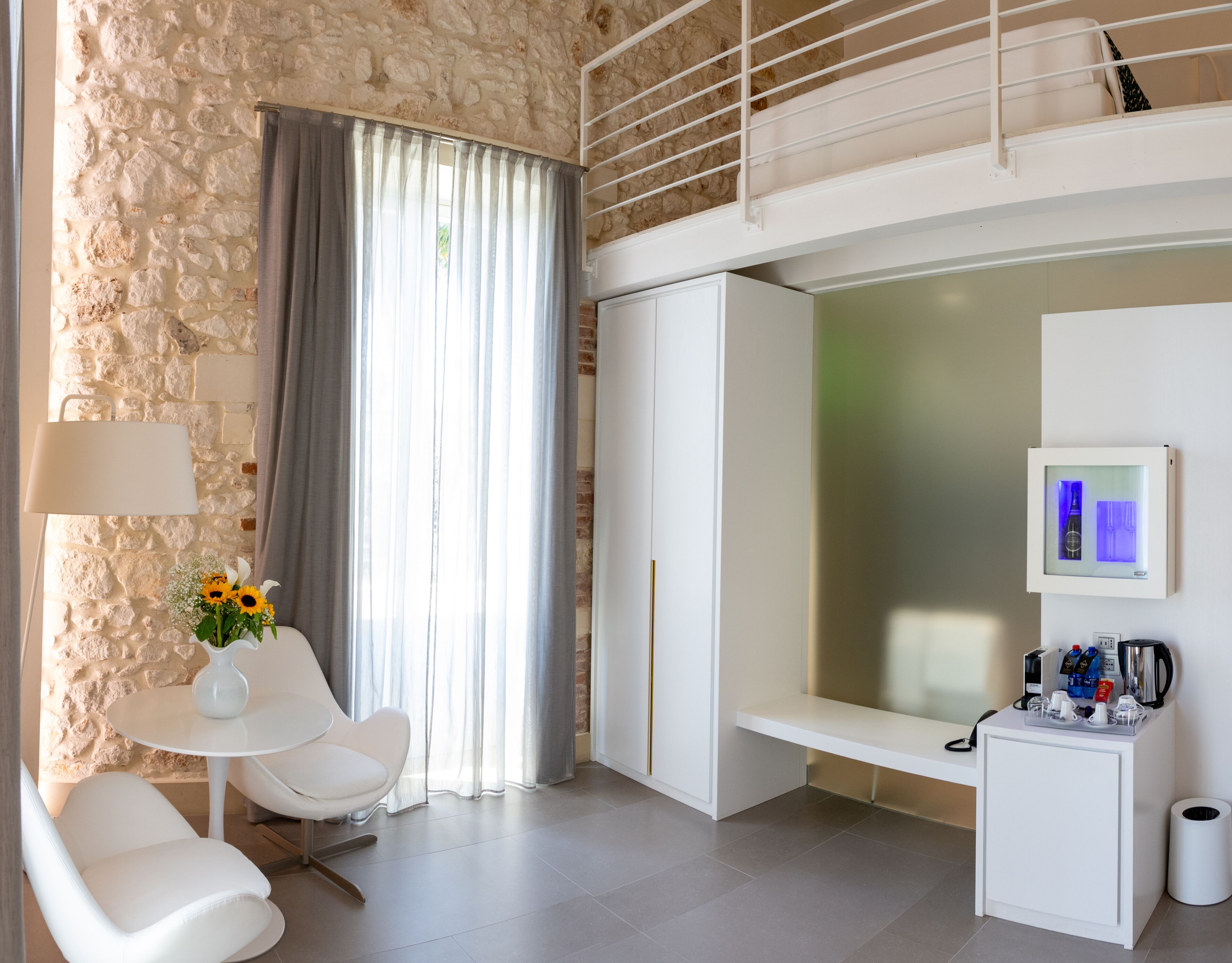 A modern hotel room with a stone wall, a balcony, a seating area with a table and chairs, a minibar, and a large window with curtains.