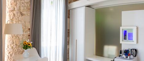 A modern hotel room with a stone wall, a balcony, a seating area with a table and chairs, a minibar, and a large window with curtains.