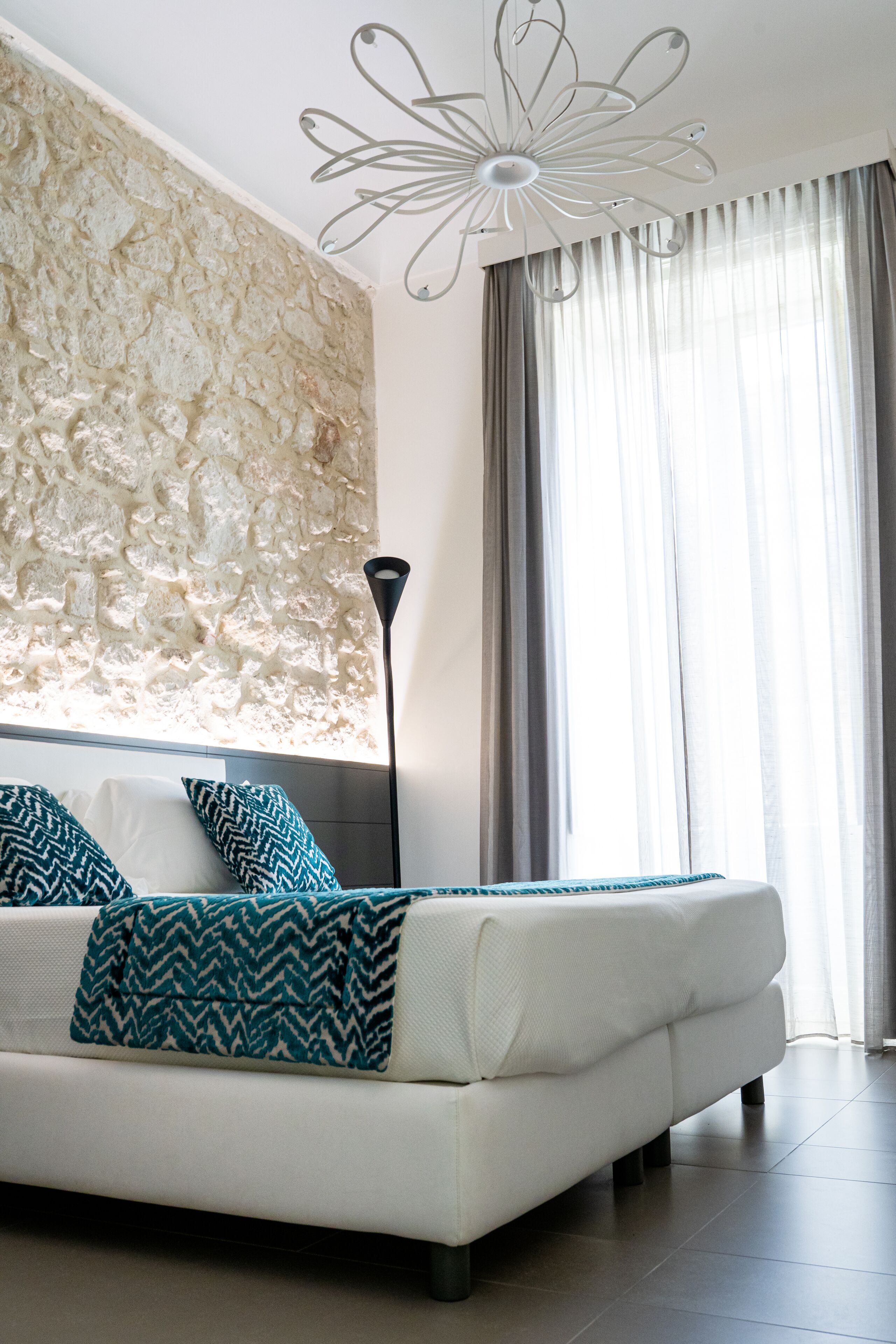 A modern bedroom with a stone accent wall, a white sofa, patterned throw pillows, a floor lamp, and a large window with curtains.