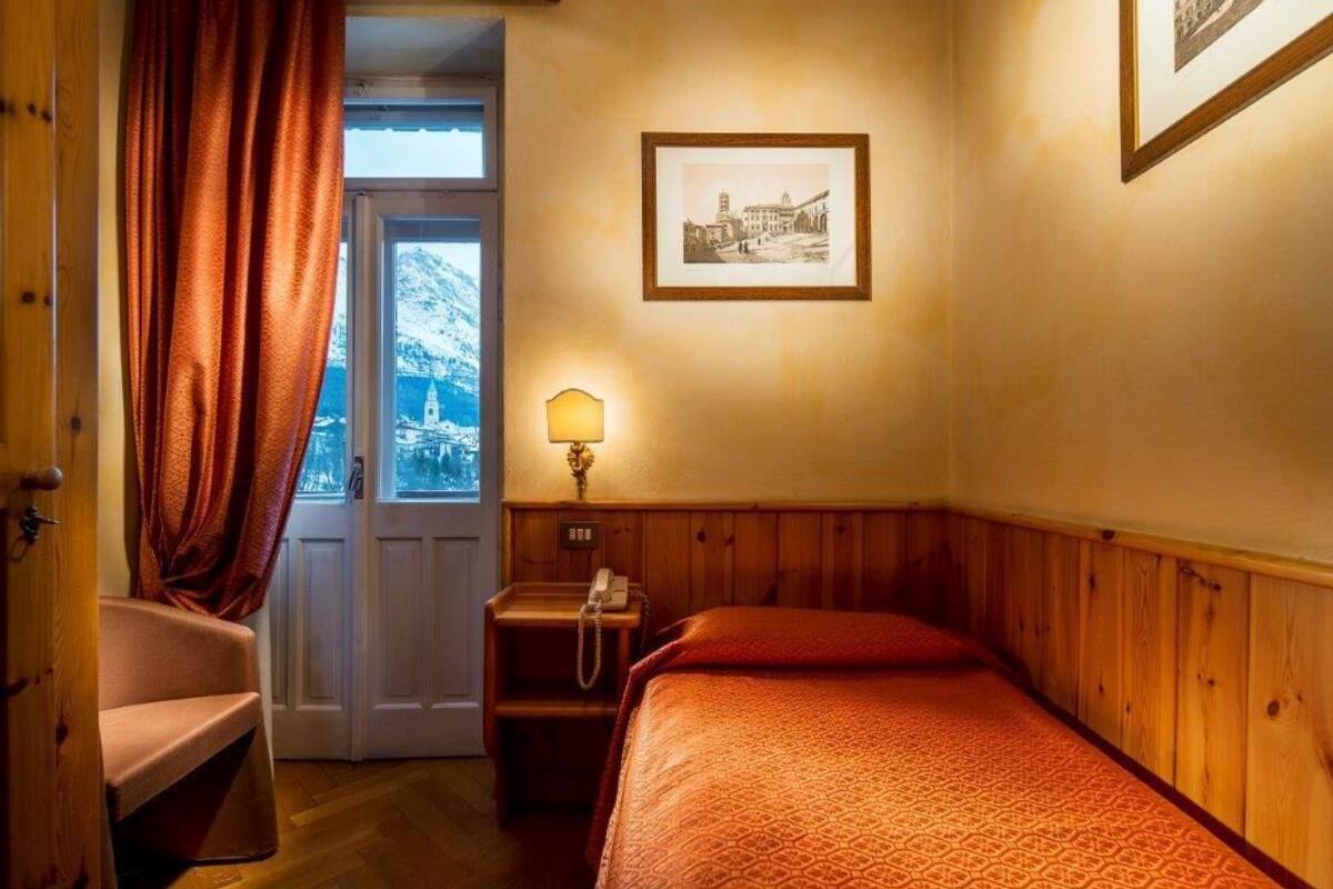 Ein Hotelzimmer mit einem hölzernen Bett, einem Sessel, einem Nachttisch mit Lampe, einem gerahmten Bild an der Wand und einem Fenster mit Vorhängen.