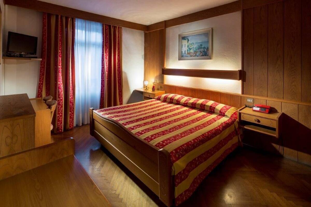 Ein Hotelzimmer mit einem hölzernen Bett, einem Schreibtisch mit Stuhl, einem Fernseher an der Wand und einem Bild über dem Bett.