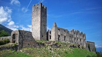 Un castello in pietra storica con una torre imponente e numerosi archi, situato su una collina erbosa sotto un cielo sereno.