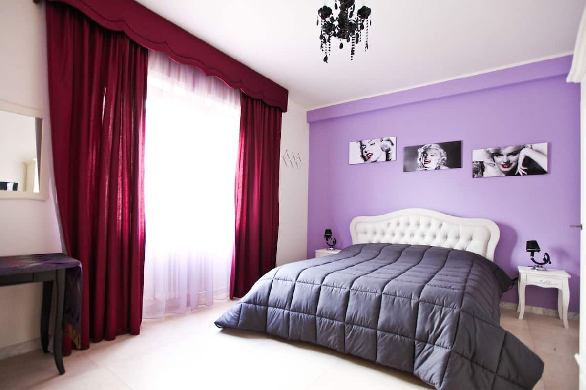 Ein Schlafzimmer mit einem großen Bett, einem Nachttisch, einer Kommode und einem Fenster mit roten Vorhängen.