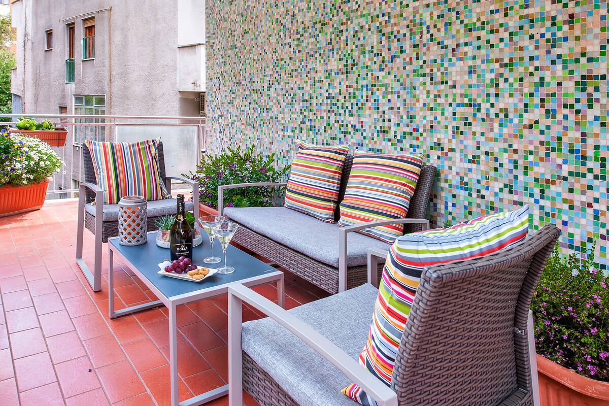 Ein Balkon mit Korbmöbeln, Fliesenboden und einer farbenfrohen Mosaikwand.