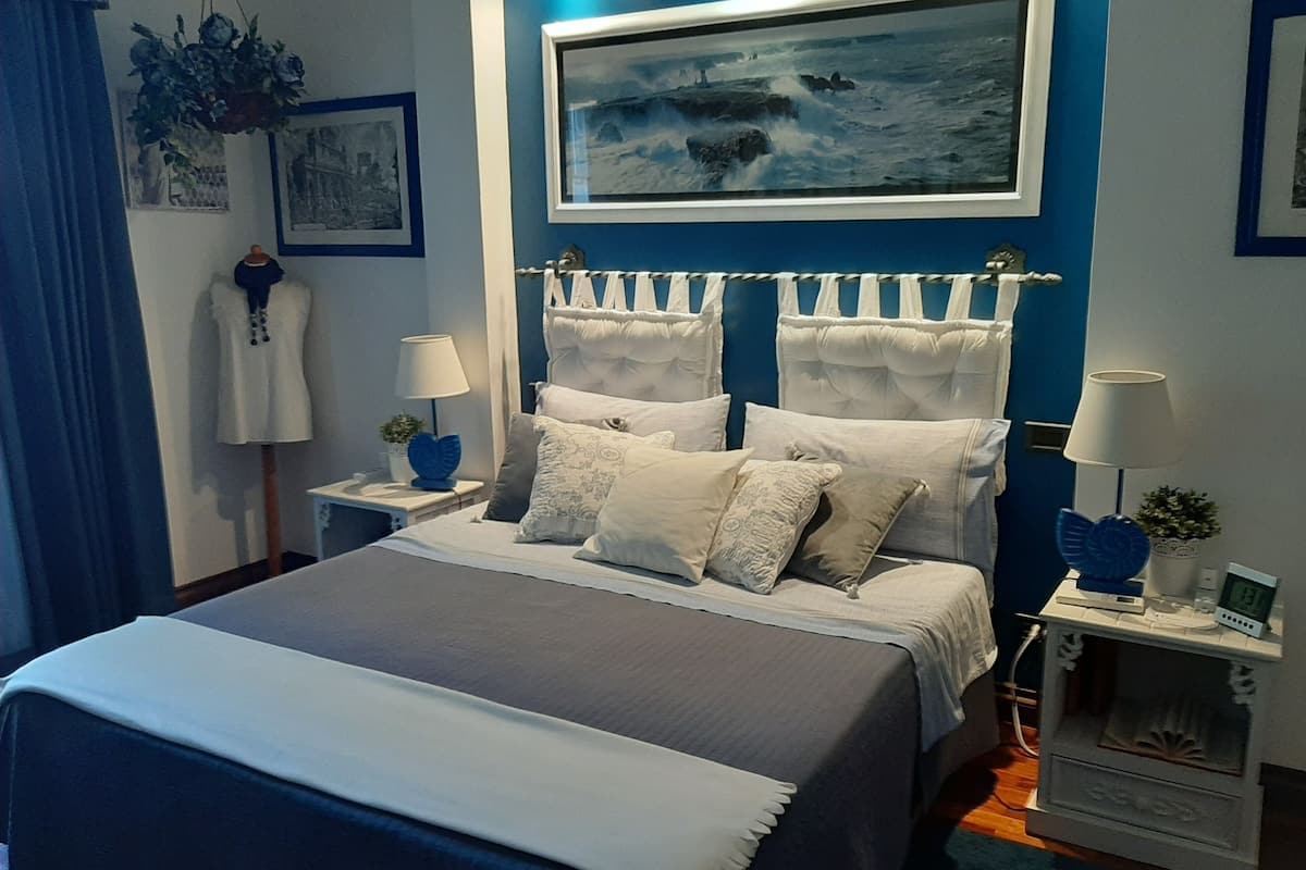 A bedroom with a bed, two bedside tables, a nightstand, a wall-mounted painting, and a blue curtain.