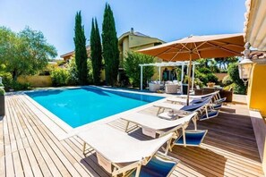 Zona junto a la piscina con sillones reclinables y sombrillas, rodeada de vegetación y un edificio al fondo.