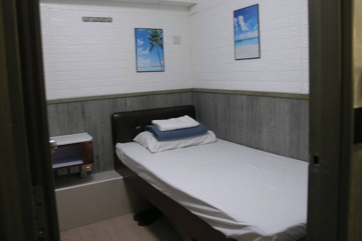Ein kleiner, schmaler Raum mit einem Einzelbett, einem Tisch und einem an der Wand befestigten Regal. Zwei gerahmte Bilder hängen an der Wand.