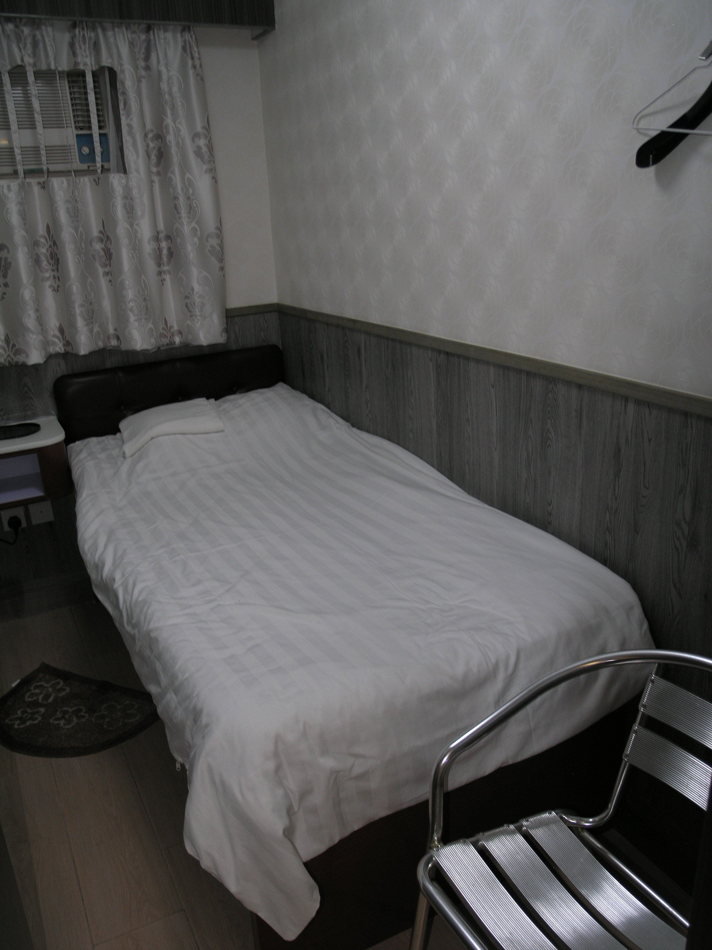 A single bed with a wooden headboard, a metal chair, a small table, and a window with a curtain.