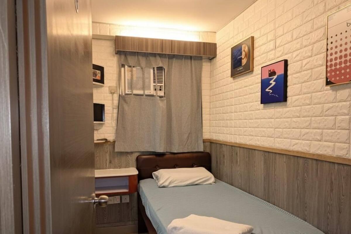 Ein kleines, aufgeräumtes Schlafzimmer mit einem Einzelbett, einem Nachttisch, einer Wand-Klimaanlage und einem Fenster mit Vorhängen.