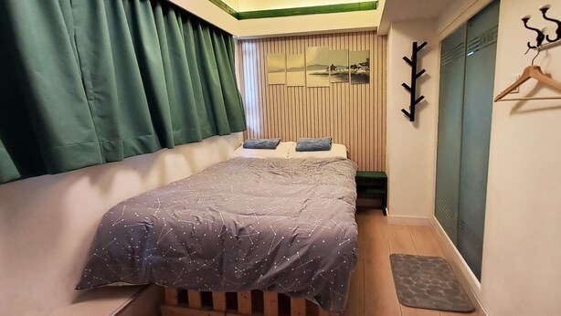 Une chambre à coucher avec un lit, une tête de lit en bois, une petite table, un porte-manteau mural et une vue sur un paysage par la fenêtre.