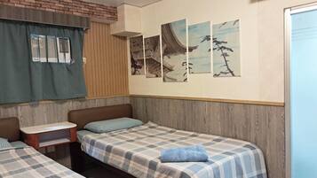 A room with two single beds, a small table, a window with blinds, and a wall with three framed pictures.
