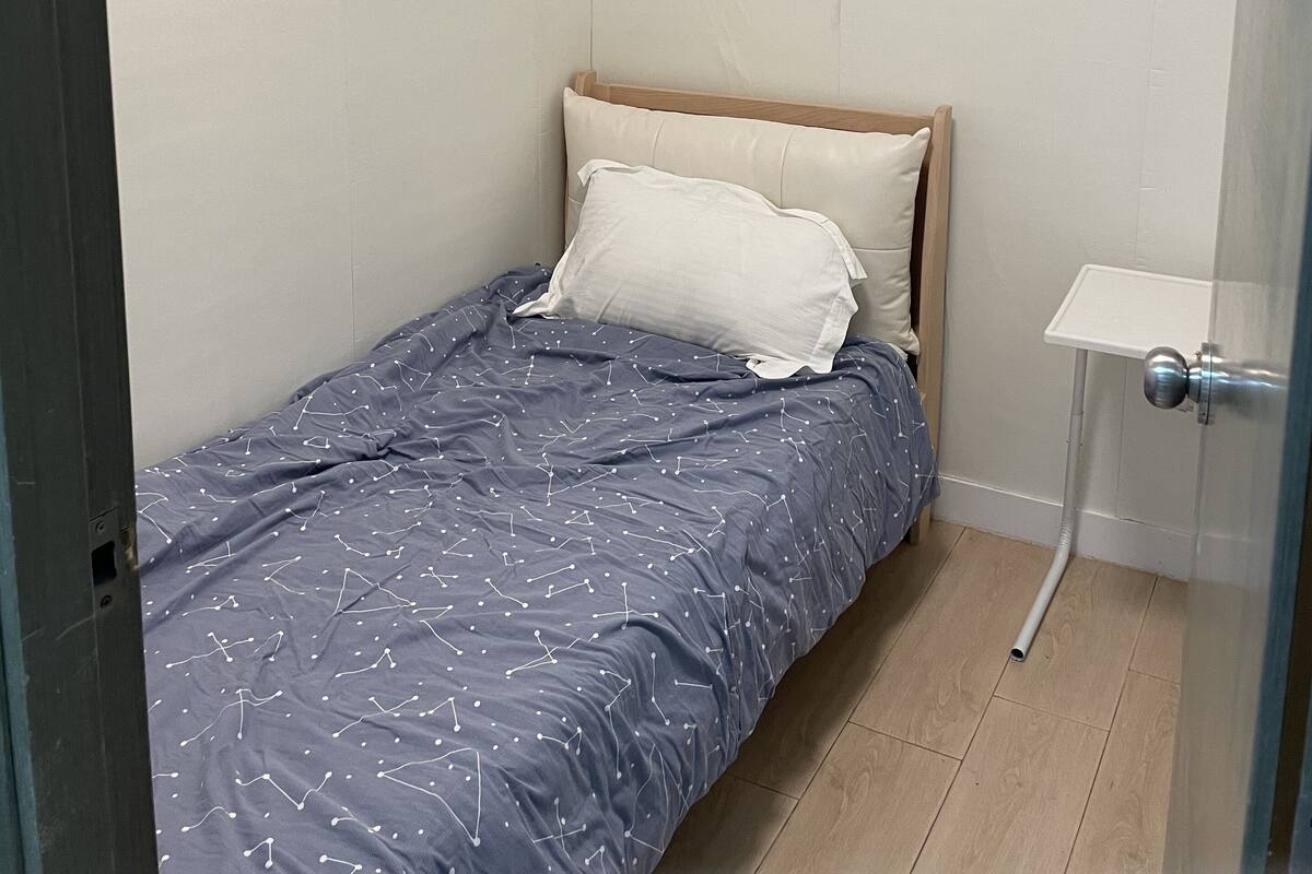 Ein kleines Zimmer mit einem Einzelbett, einem hölzernen Kopfteil, einem weißen Kopfkissen und einer blauen Bettdecke mit weißen Mustern.