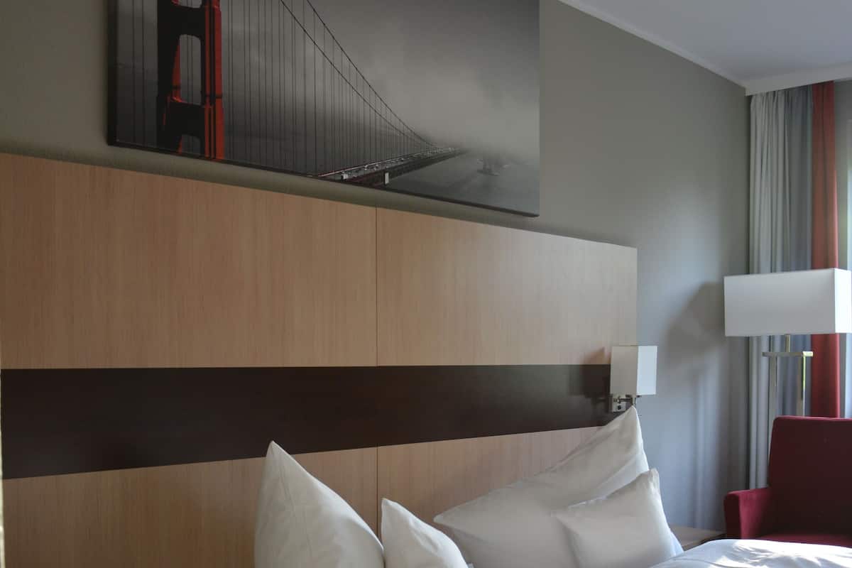 Quarto de hotel com uma cama grande, cabeceira com painéis de madeira horizontais, uma cadeira vermelha, um abajur e um quadro da Ponte Golden Gate.