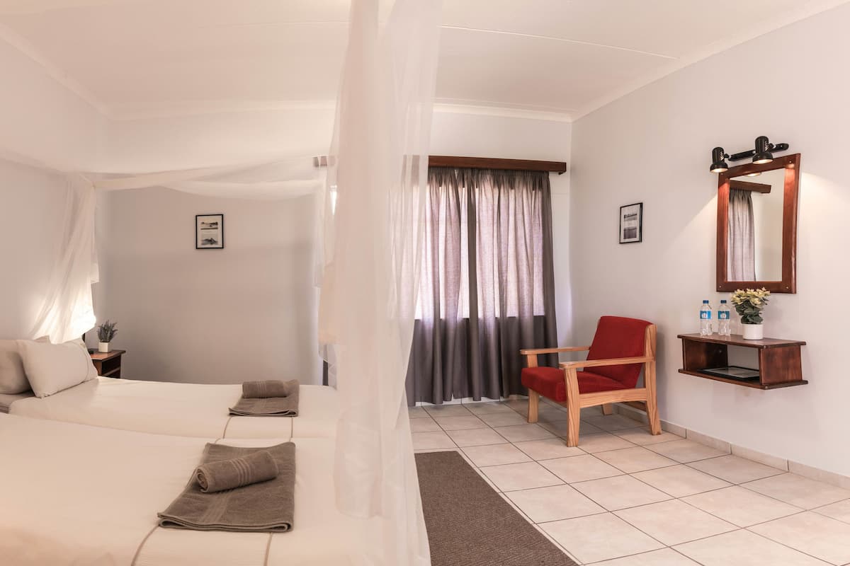 Hotellihuone, jossa on kaksi sänkyä, punainen tuoli, pieni pöytä, jossa on kasvi, ja peili seinällä.