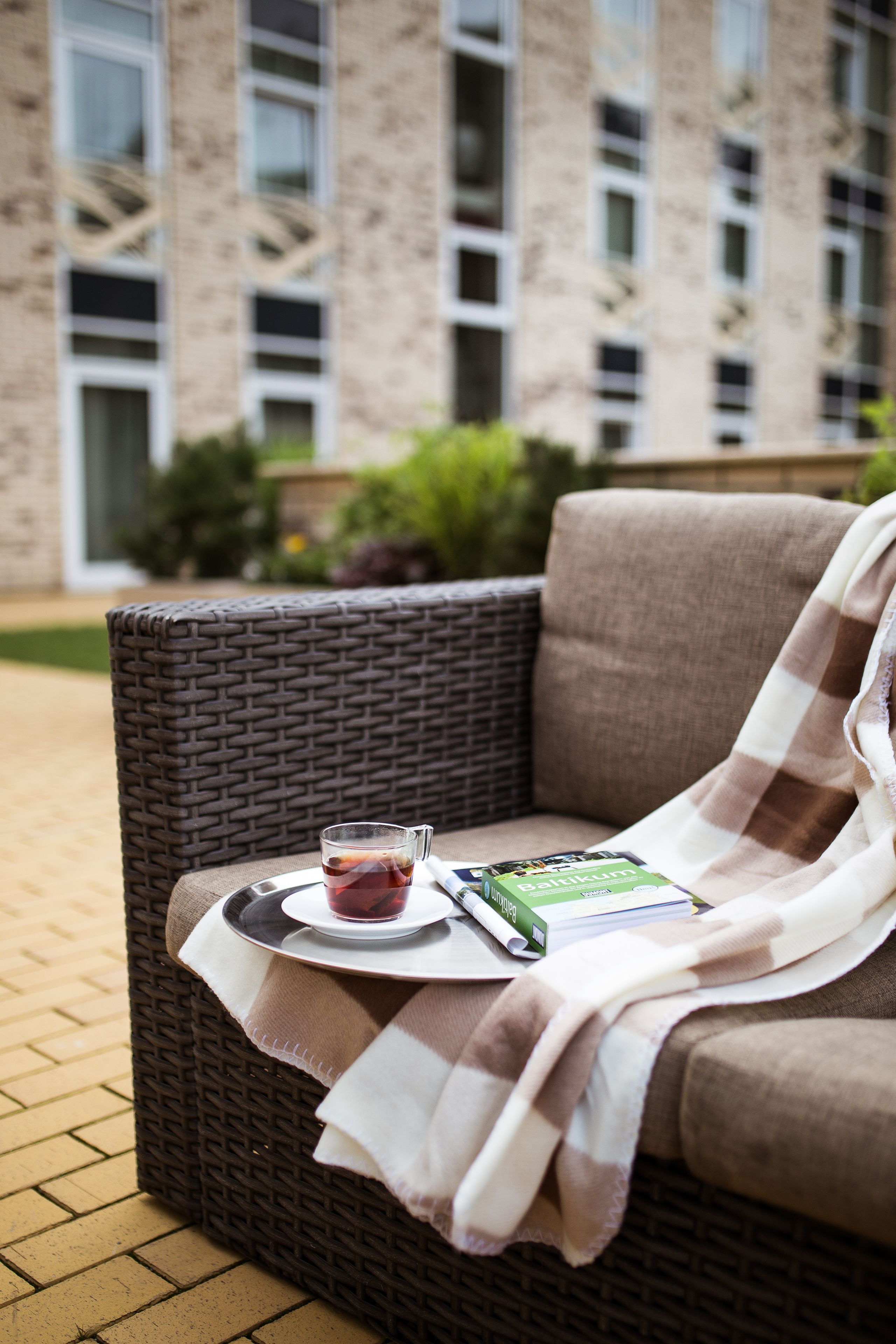 A wicker chair with a tray holding a glass of beverage and a book, set on a brick-paved area with a building in the background.