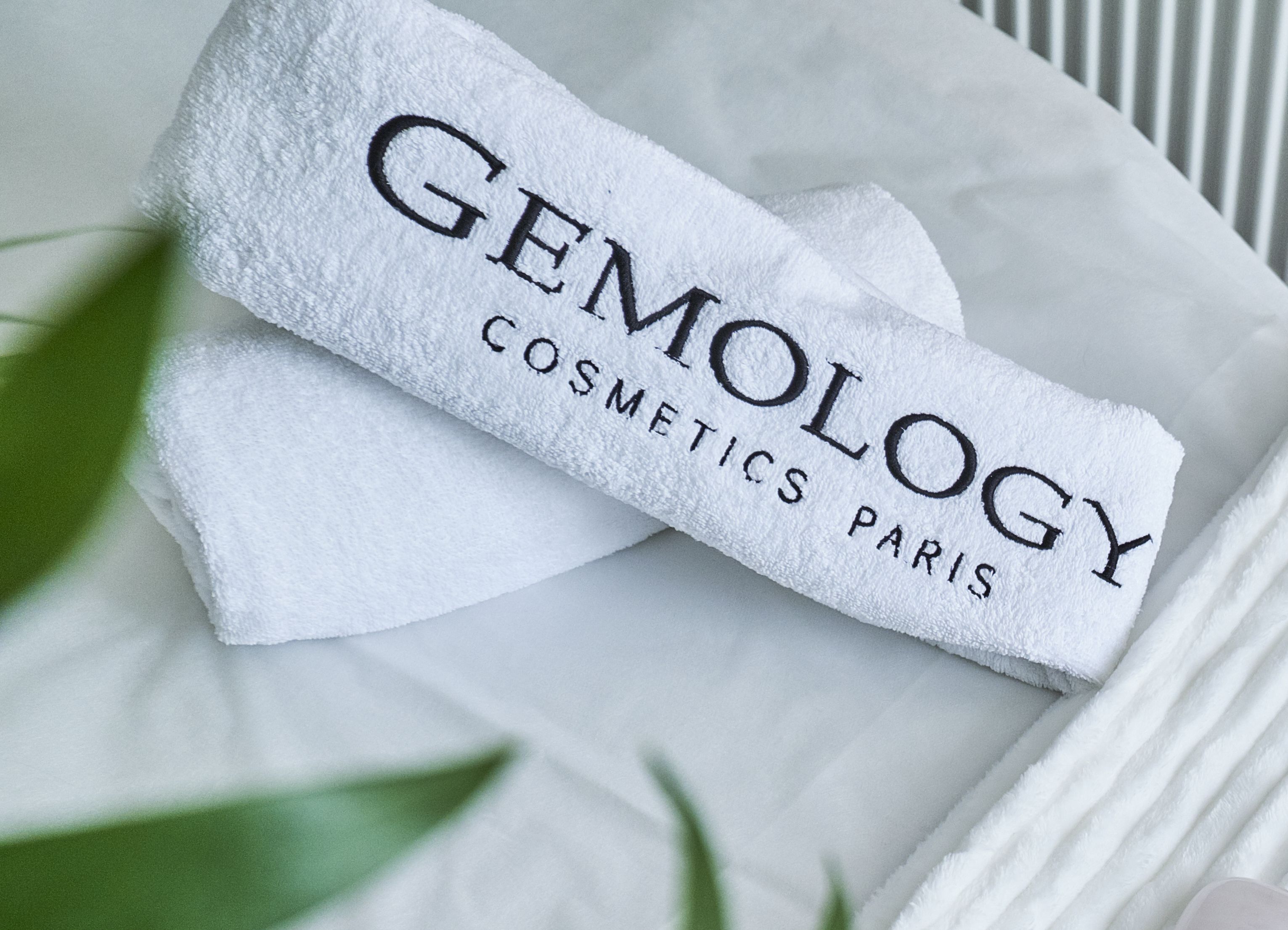 A white towel with "GEMOLOGY COSMETICS PARIS" printed on it, placed on a white surface with green leaves in the background.