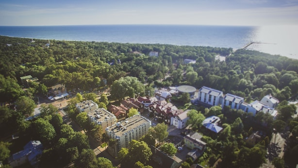 An aerial view of a coastal town with dense greenery, a mix of residential and commercial buildings, and a visible body of water. - Grand Baltic Dunes (Palanga)