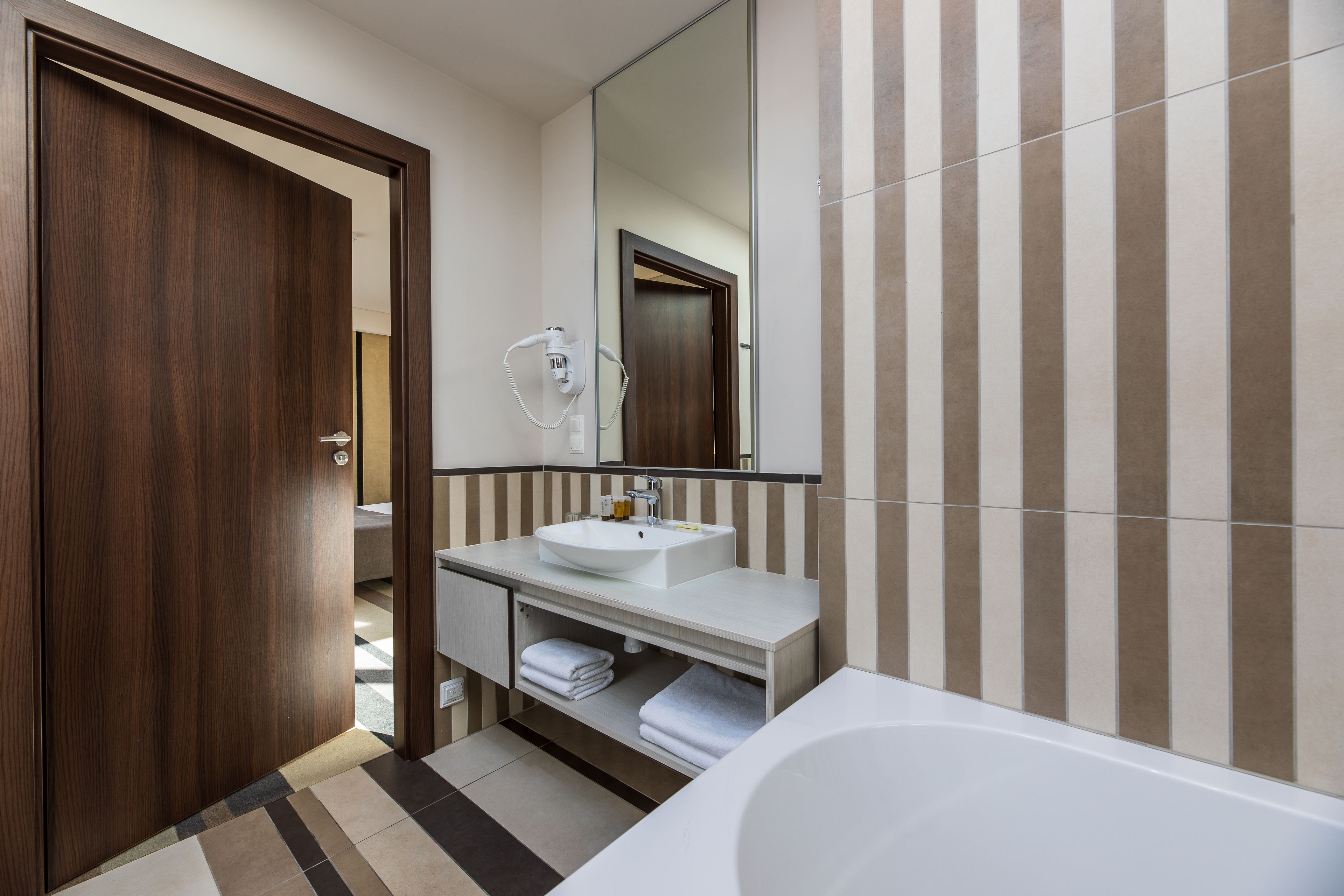 Un baño moderno con puerta de madera, un lavabo blanco, un espejo y baldosas a rayas.