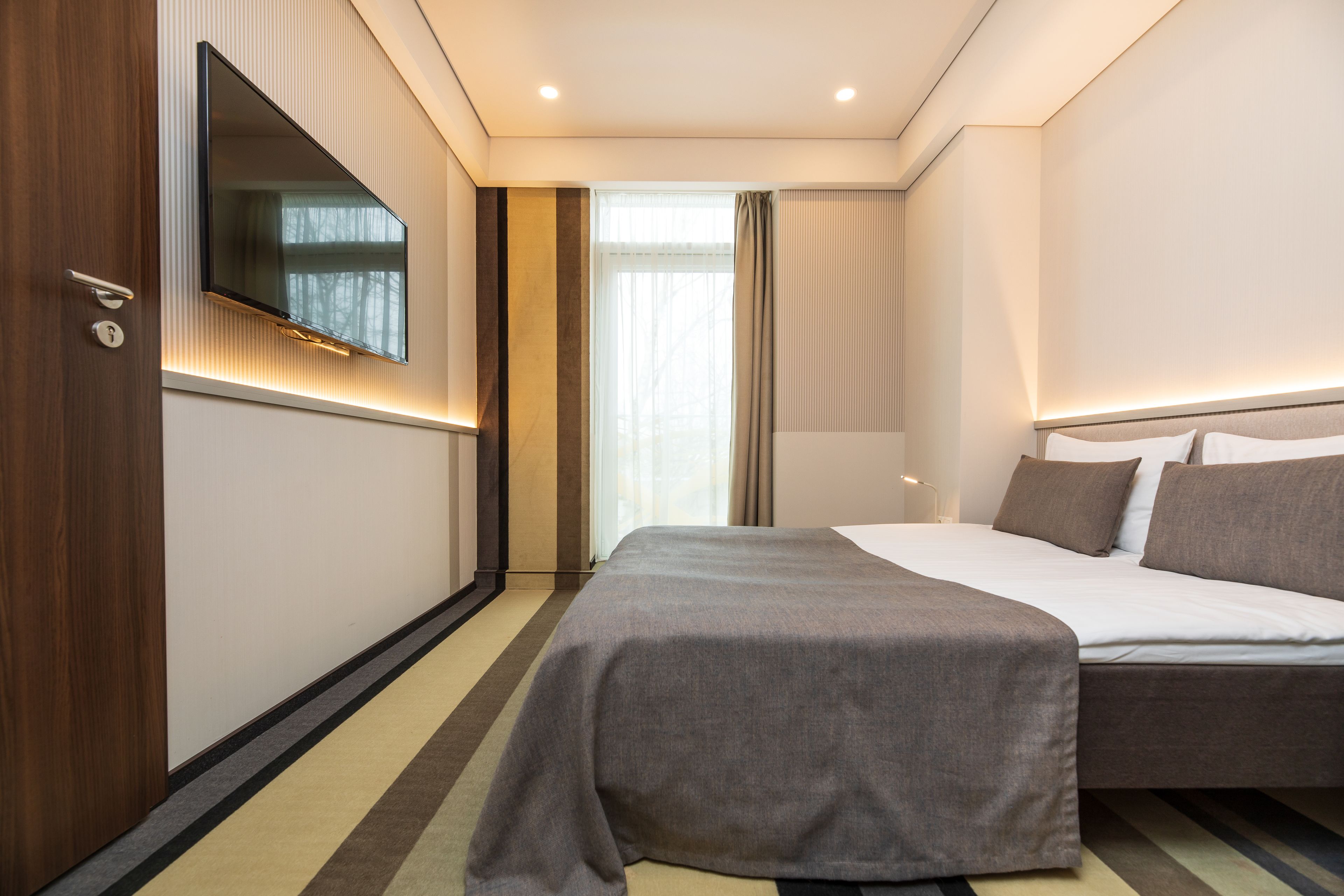 A hotel room with a bed, a TV mounted on the wall, a window with curtains, and a door with a handle.