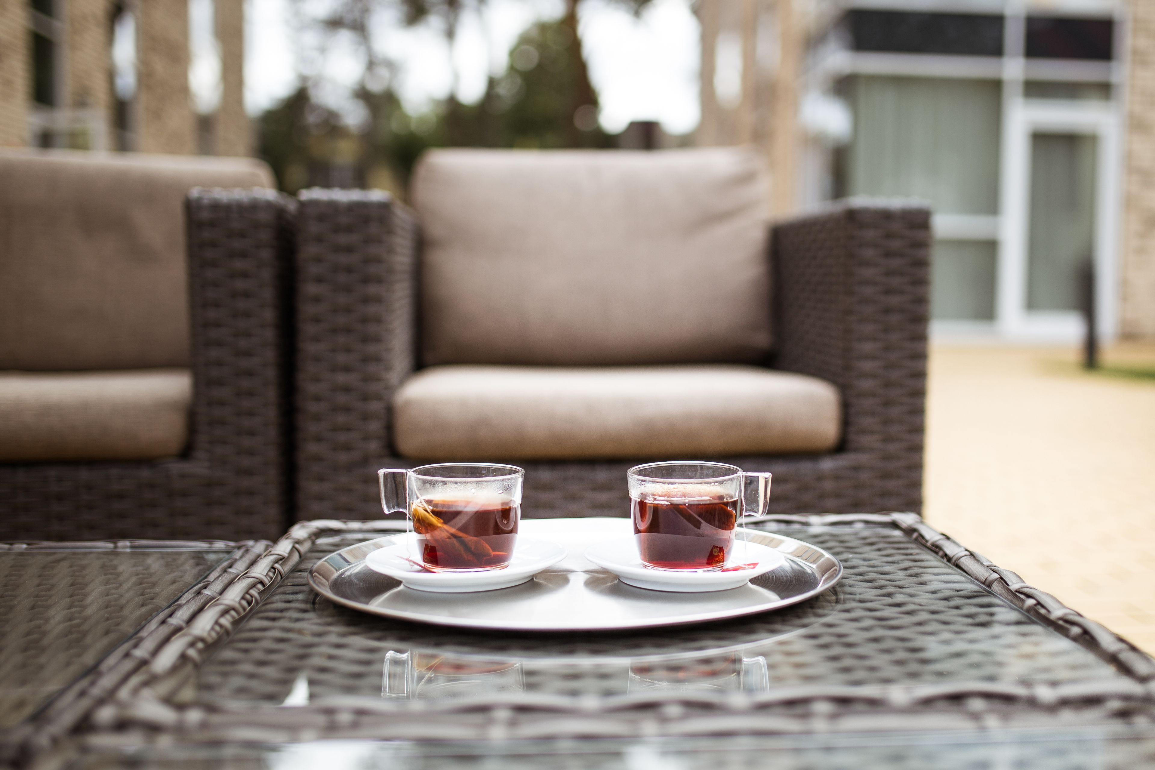 Two cups of tea on a tray on a glass table with wicker chairs in the background.