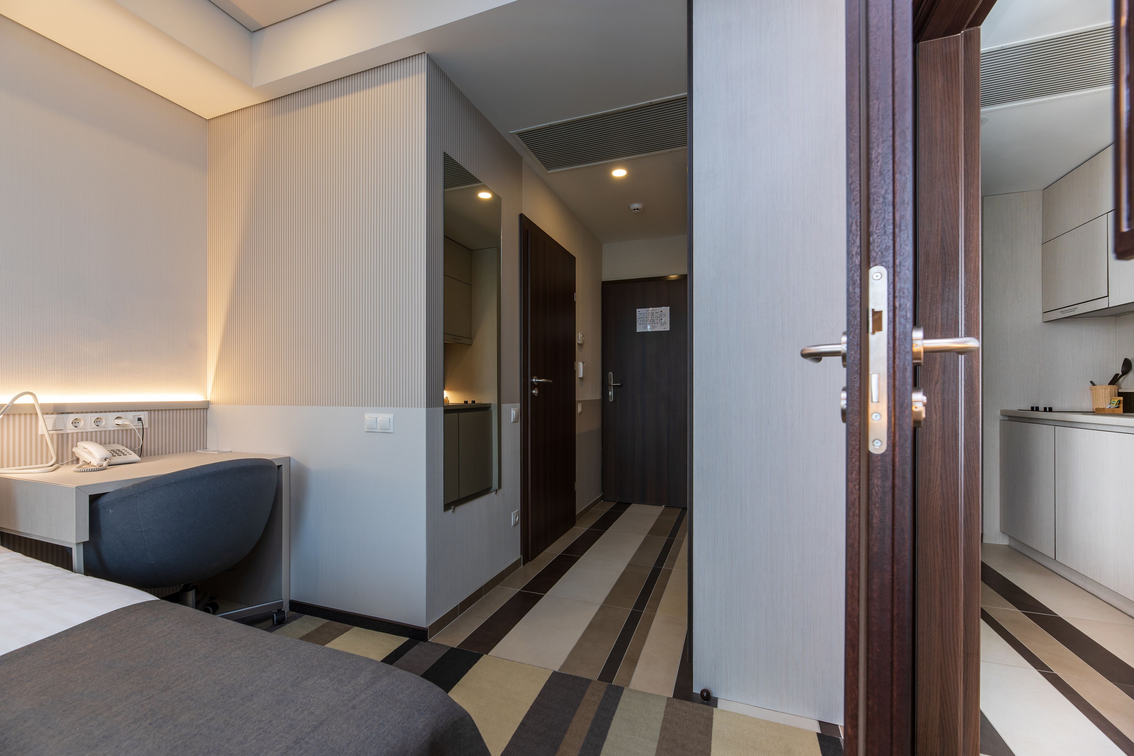 A hotel room with a bed, bedside table, lamp, and a view of the hallway leading to the kitchen.