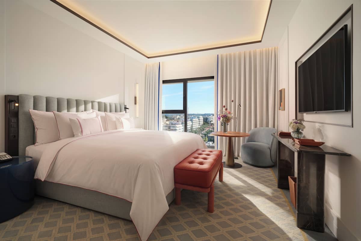 Habitación de hotel moderna con una cama grande, zona de estar, una pequeña mesa con flores, televisión y vista a la ciudad.