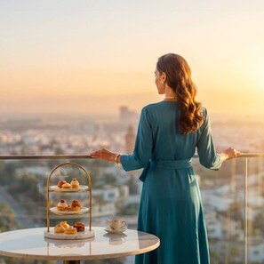 Una persona está de pie en un balcón con vistas a una ciudad, con un soporte escalonado para postres y una taza sobre una mesa.