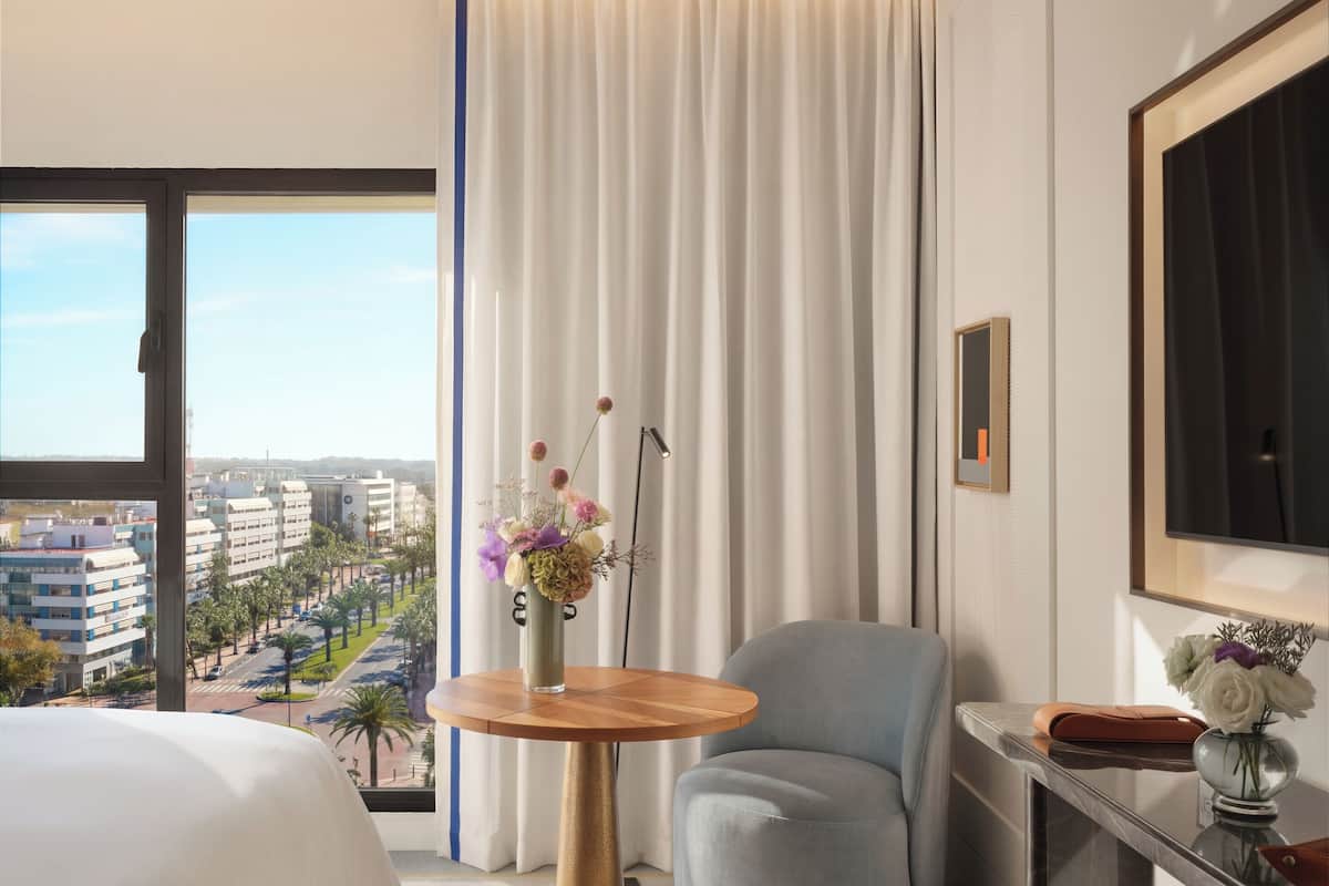 Habitación de hotel con una cama grande, una mesa redonda de madera con un jarrón de flores, un sillón gris, un escritorio con un espejo y vistas a un paisaje urbano a través de amplias ventanas.